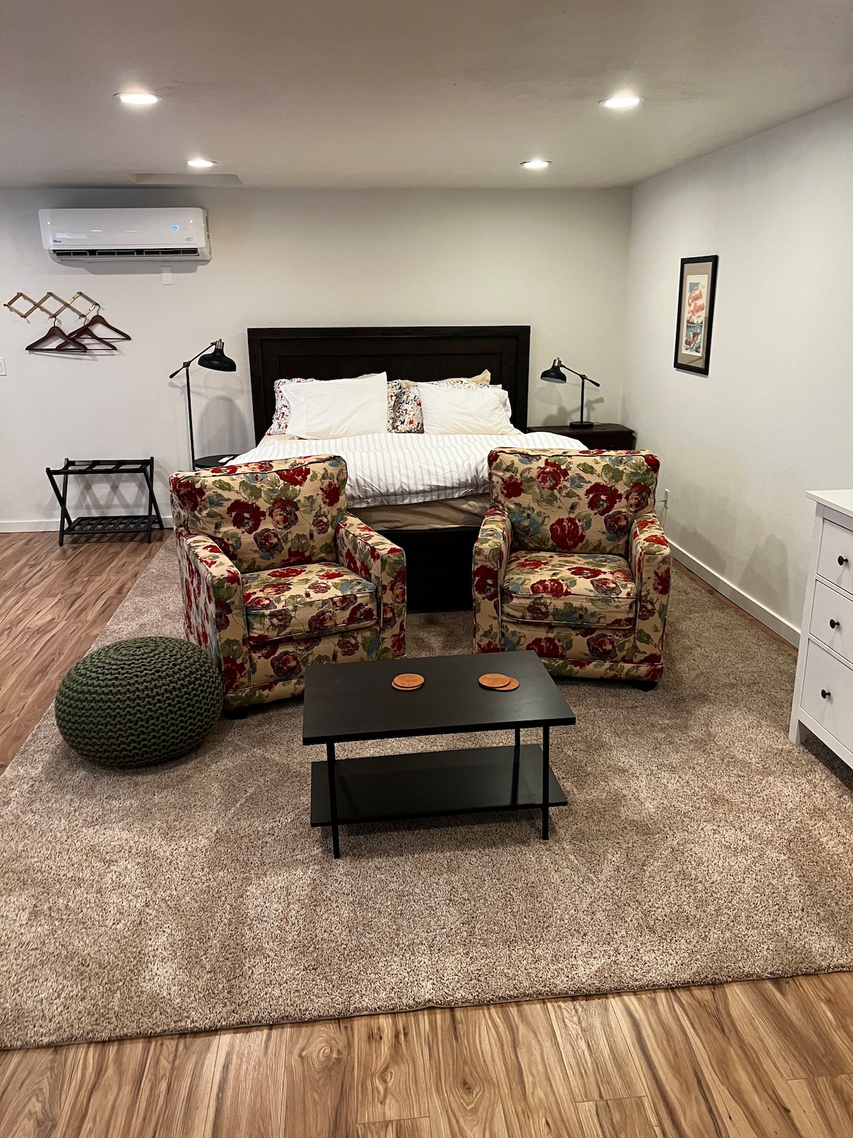 A spacious studio features a cozy sleeping area with a king bed, complemented by stylish bedding. Two floral upholstered chairs are arranged next to a small black table, and a green pouf is placed nearby. Soft lighting is provided by lamps beside the bed.