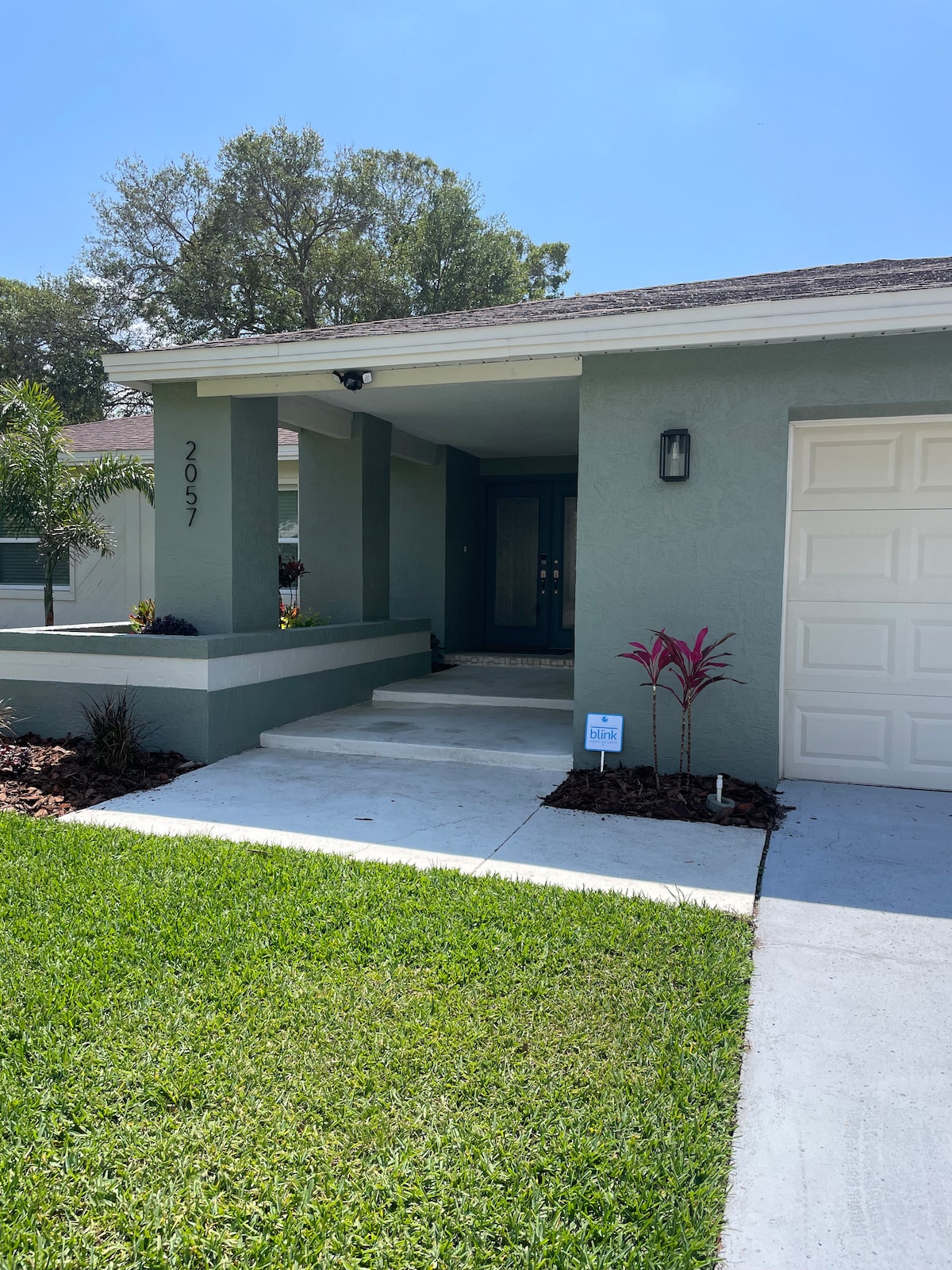 A welcoming facade of the house features a covered entrance with a porch, flanked by landscaped greenery. The smooth concrete pathway leads to a dark front door, and a single-car garage is visible on the right, surrounded by well-maintained grass.