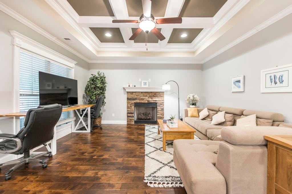 A spacious living area is illuminated by natural light from large windows. A comfortable sectional sofa faces a TV mounted on a stand, while a decorative stone fireplace provides a focal point. A wooden coffee table and a desk with a chair complement the warm wooden flooring.