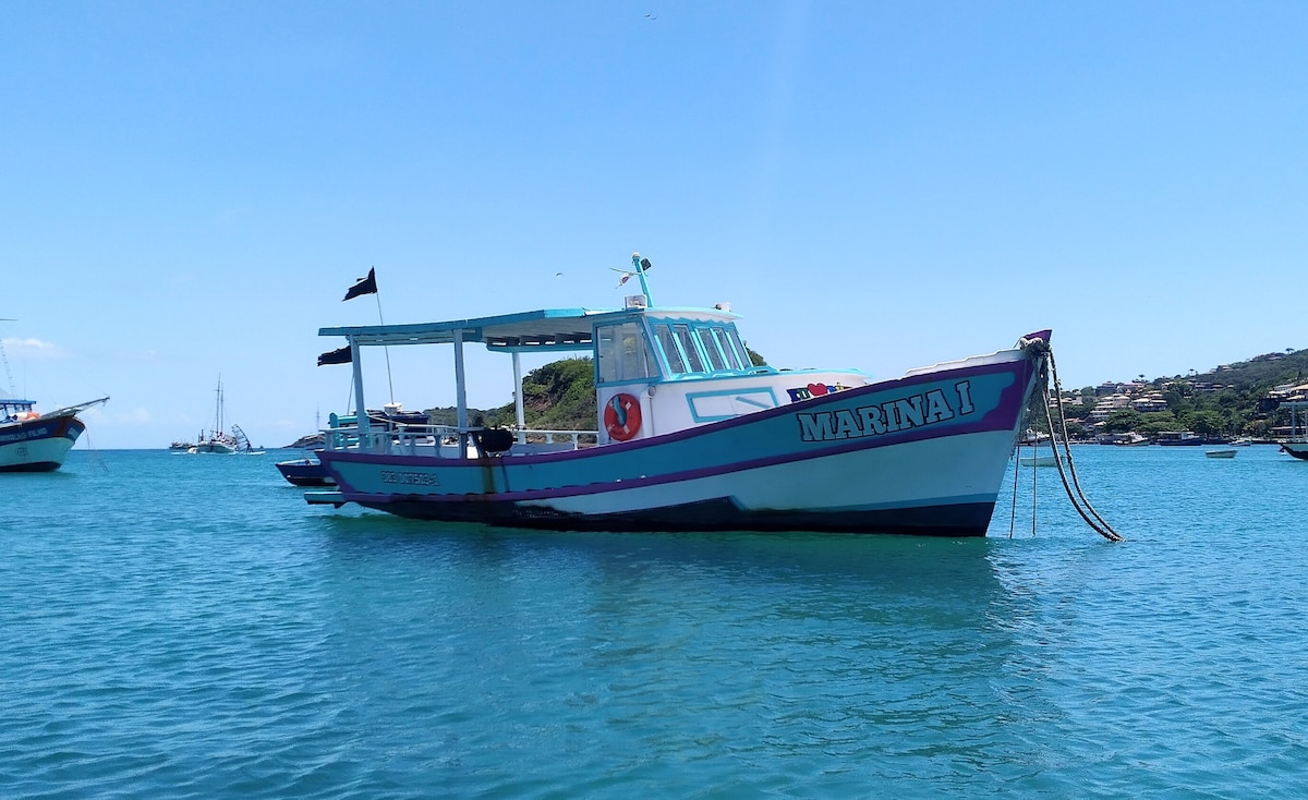 A colorful boat named 'Marinai' is anchored in calm blue waters, with a bright blue sky overhead. The boat features a spacious deck and is surrounded by other boats and lush green hills in the background.