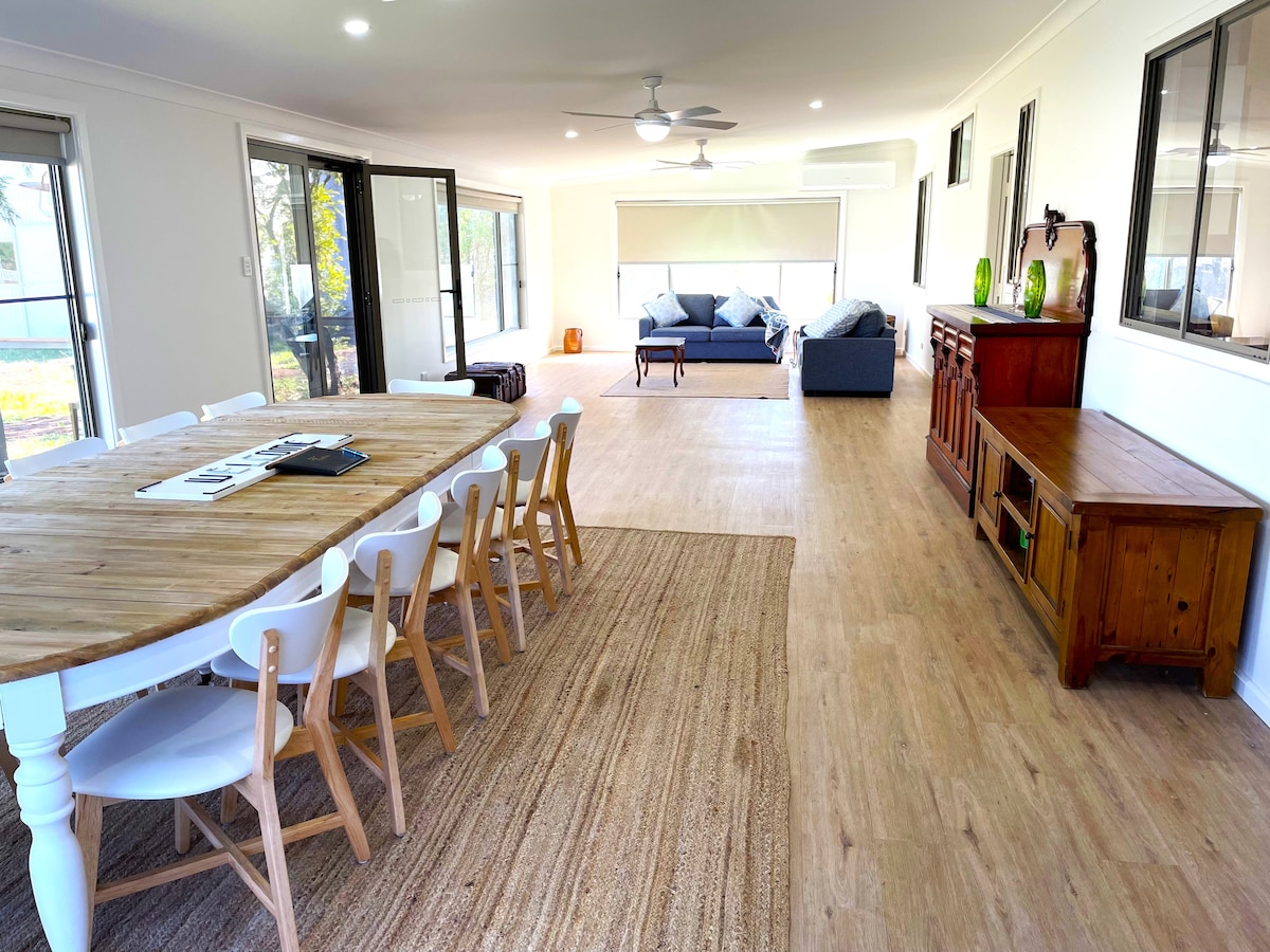 A spacious family room is showcased, featuring a large round dining table with white chairs. In the background, a comfortable seating area with soft couches is visible. Ample natural light floods the space through multiple windows, enhancing the open layout.