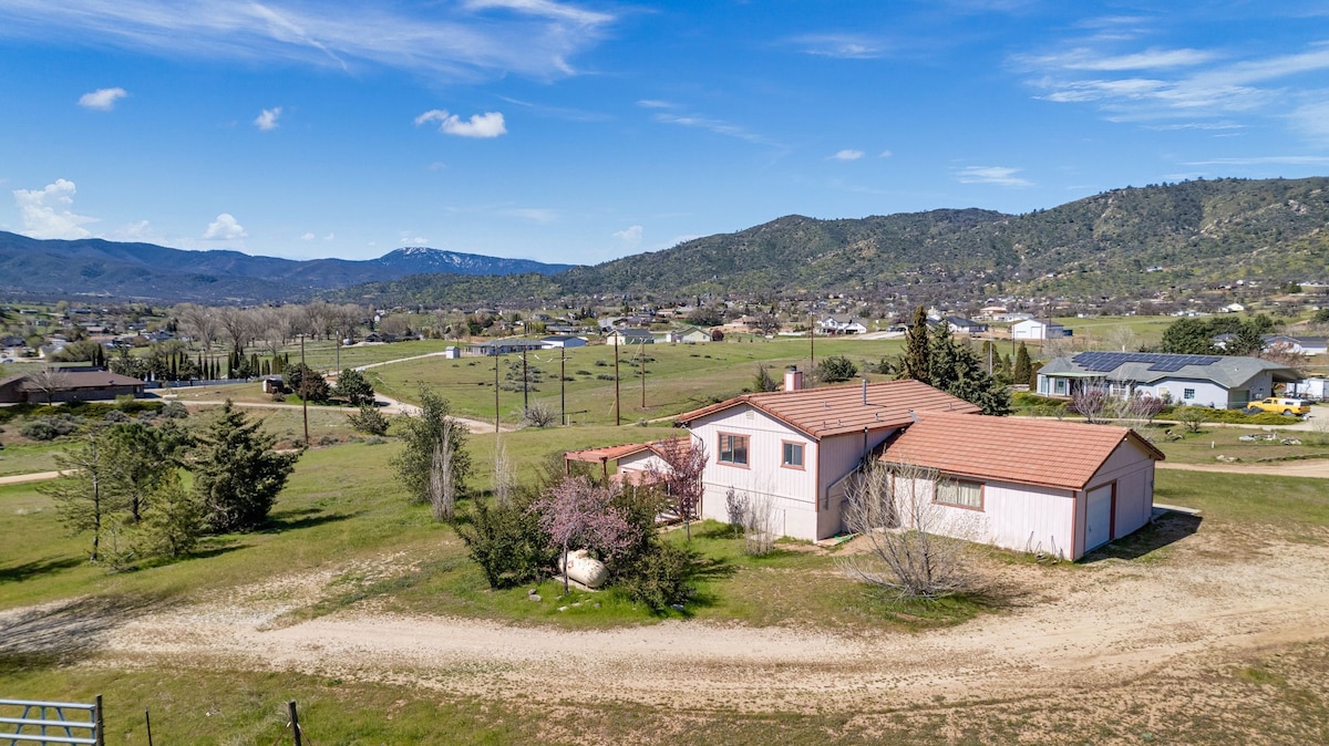 An aerial view captures a single-story home surrounded by expansive green fields and gentle hills. The structure features a red-tiled roof and appears complemented by blooming trees. Nearby, the quiet landscape extends with scattered homes, showcasing the rural charm of the Oak Knolls area.