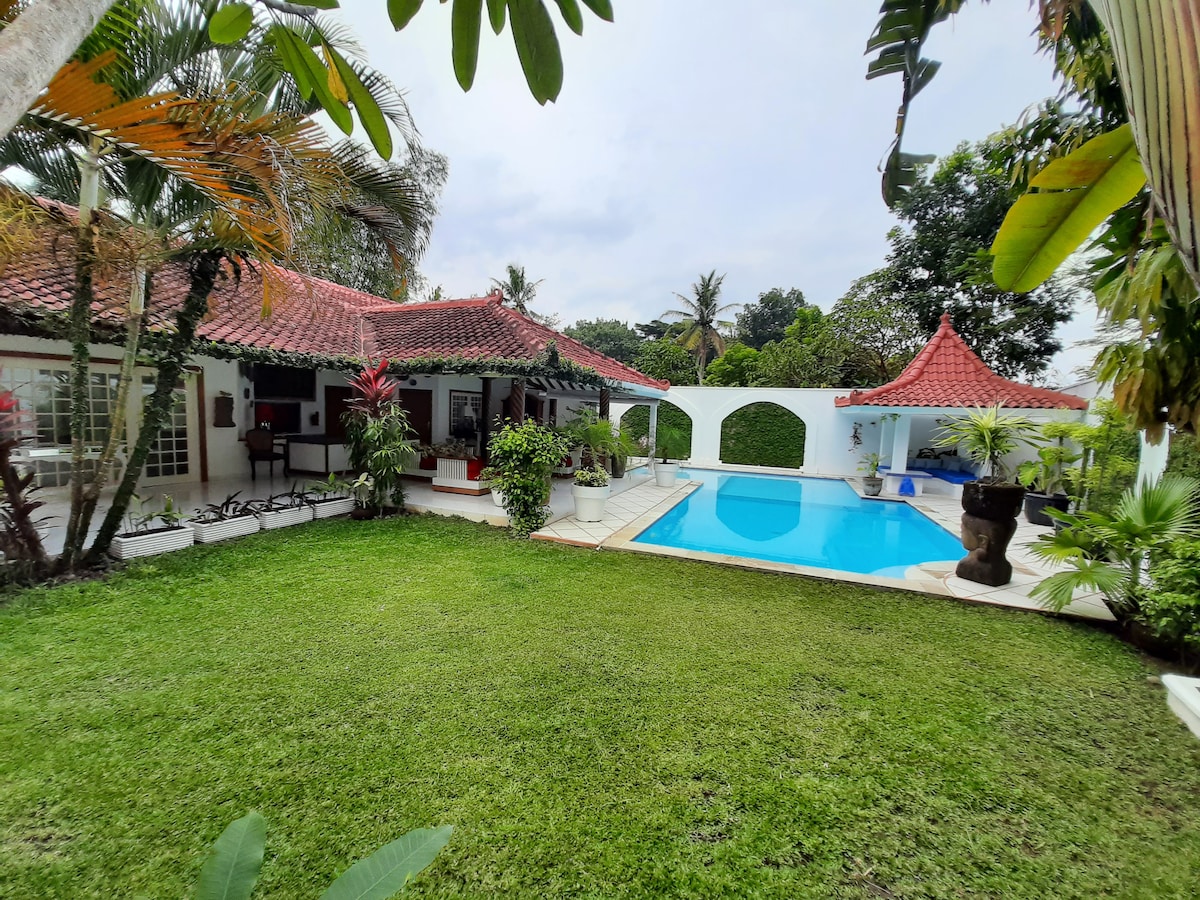 The villa's exterior features a well-maintained garden with lush greenery surrounding a private swimming pool. The traditional architecture is highlighted by a red-tiled roof and a gazebo, enhancing the inviting outdoor space. Tropical plants add character to the serene atmosphere.