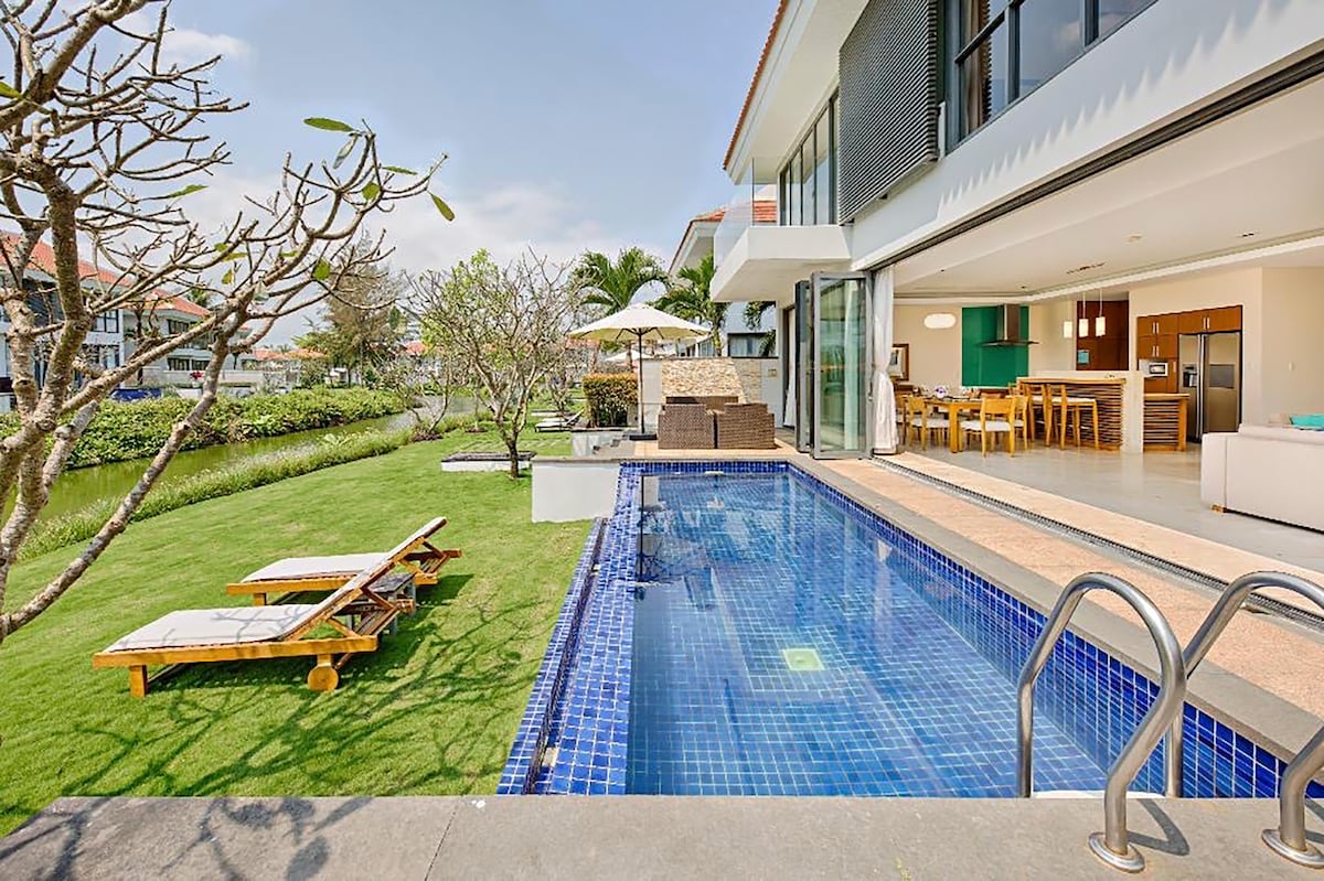A spacious outdoor area features a private pool with steps leading in, bordered by lounge chairs. A dining area is visible under a parasol, alongside the grassy landscape and tropical foliage, offering a relaxed atmosphere adjacent to the villa's expansive glass walls.