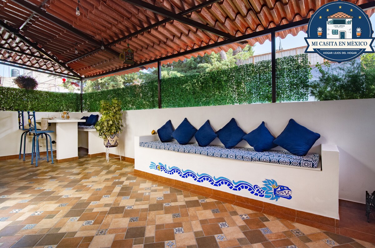 A private terrace features a cozy seating area with blue cushions arranged along a white wall. A small table is visible in the background, complemented by green plants and decorative tiles on the floor, creating an inviting space for relaxation.