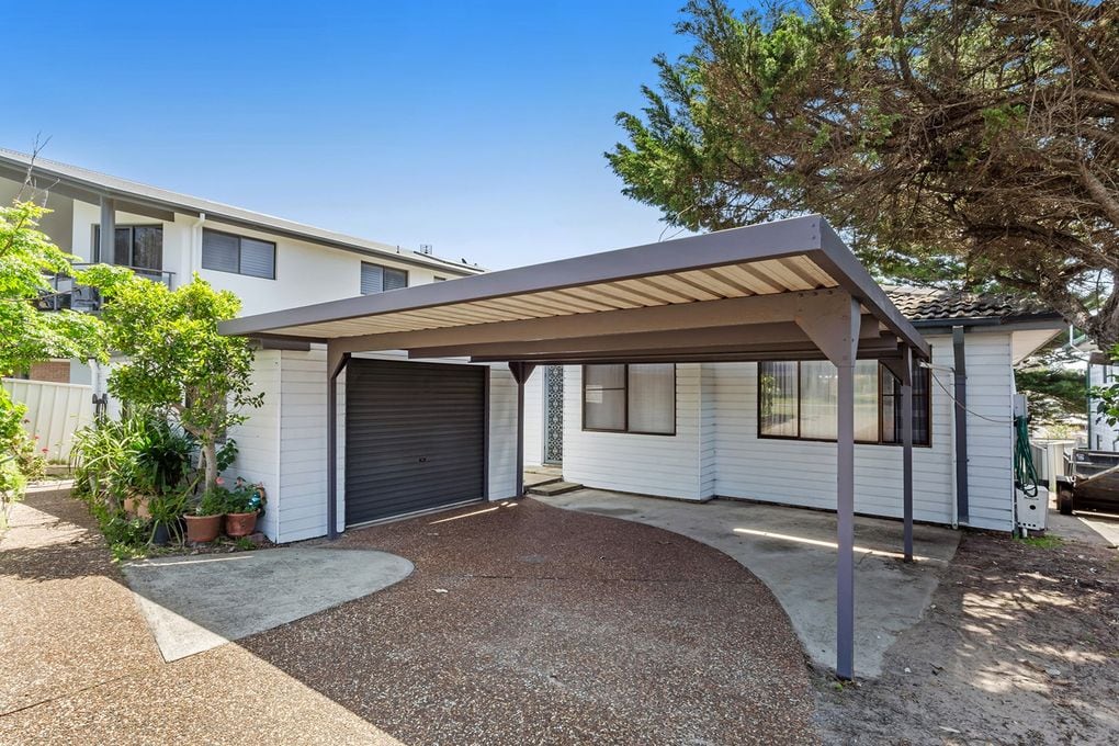 A single-story house is depicted with a covered carport, allowing for shaded vehicle access. The exterior features a combination of light-colored walls and dark trim. Adjacent greenery is visible, including potted plants and trees, enhancing the welcoming appearance of the entrance.