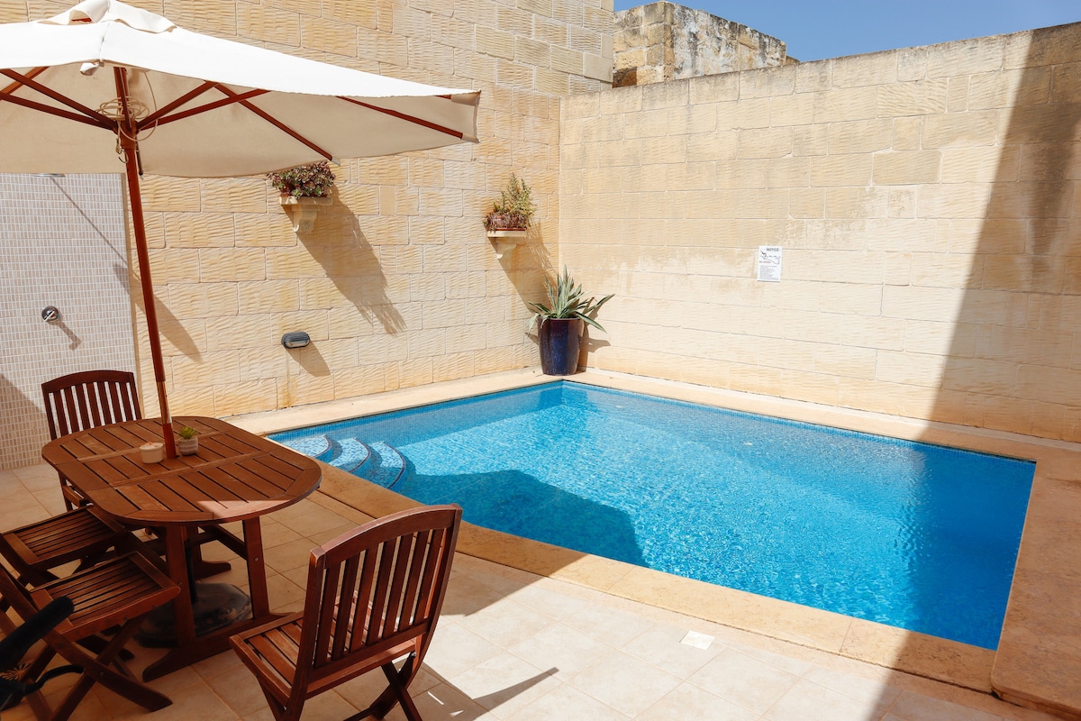 A private courtyard features a rectangular pool surrounded by sunlit stone walls. A wooden dining table with matching chairs is positioned nearby, and a large umbrella provides shade. Potted plants are placed along the walls, adding greenery to the tranquil space.