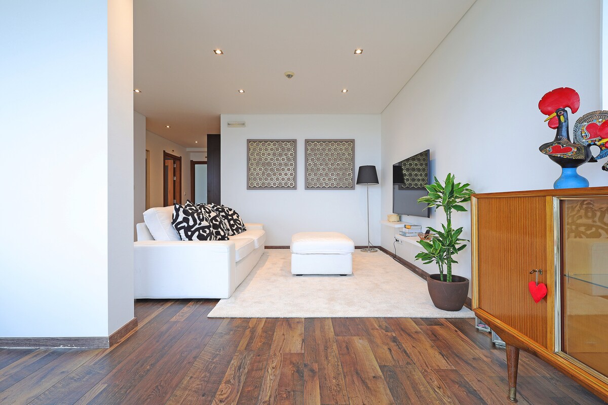 The spacious living area features a white sectional sofa with patterned throw pillows and a matching ottoman. A wooden side table holds a decorative plant. Wall art is displayed above the TV unit, enhancing the room's modern aesthetic. Wooden flooring adds warmth to the space.