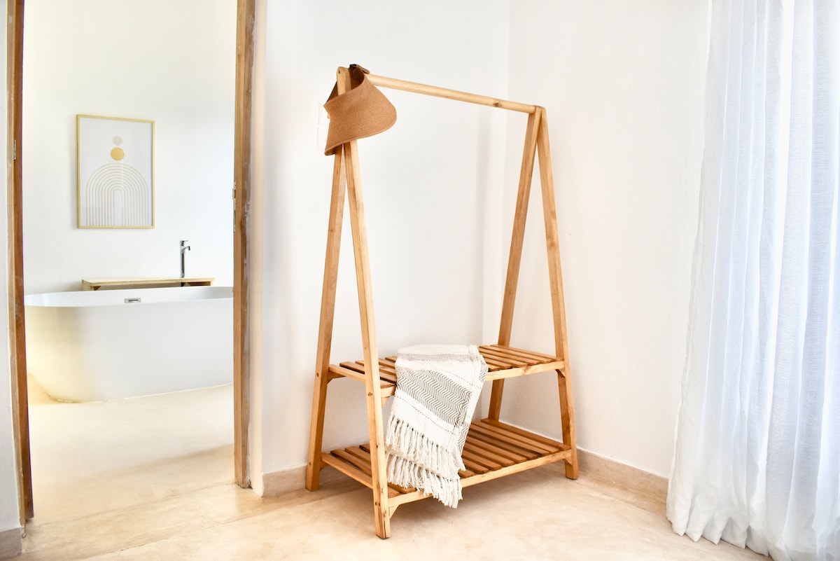 A wooden clothing rack stands in the room, featuring two levels for storage. A rolled towel is neatly placed on the lower shelf, while a hat hangs from the top rail. Sunlight brightens the space, complemented by light-colored curtains.