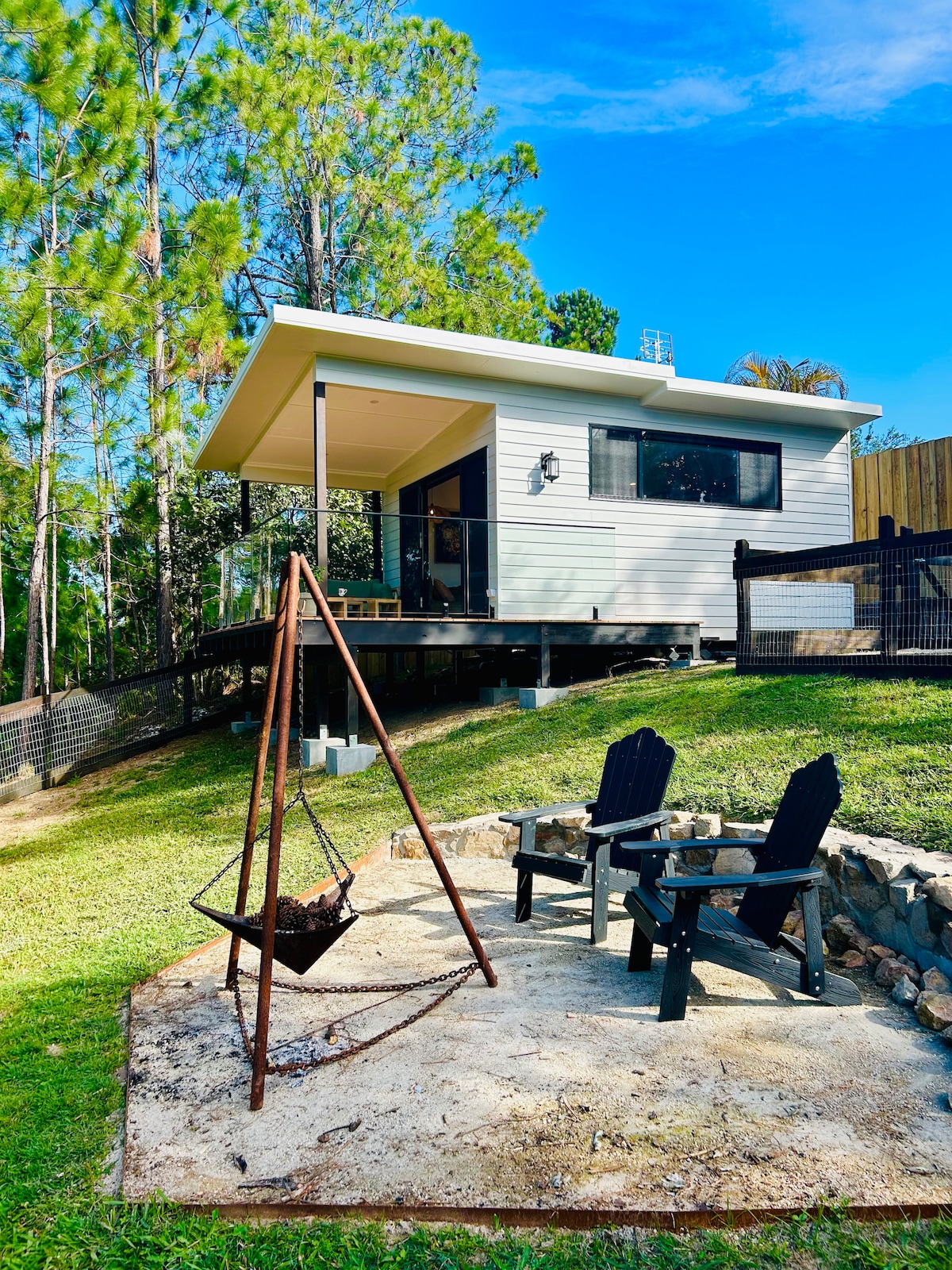 An eco cabin is set against a backdrop of tall pine trees, featuring a covered deck and large windows. A rustic fire pit is framed by two comfortable chairs, positioned on a stone patio that enhances the outdoor living space.