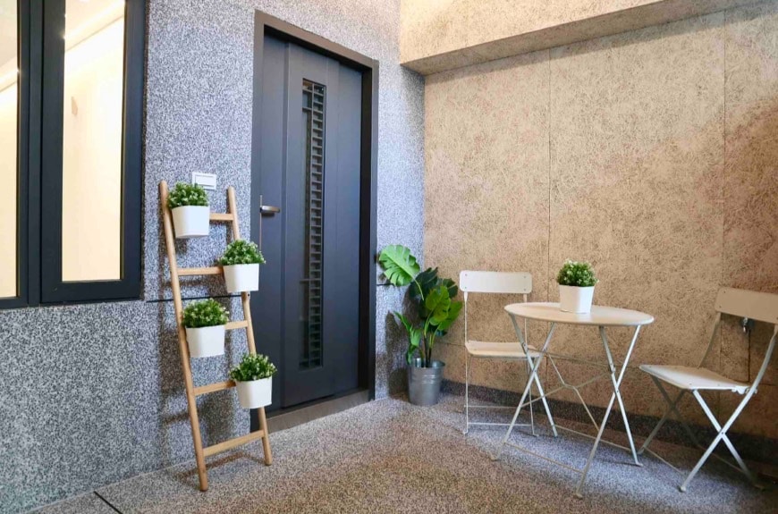 A small outdoor patio area is presented, featuring a two-person bistro table and two folding chairs. A decorative ladder holds three potted plants, and a single pot stands on the ground nearby. The entrance door is framed by minimalistic design elements.