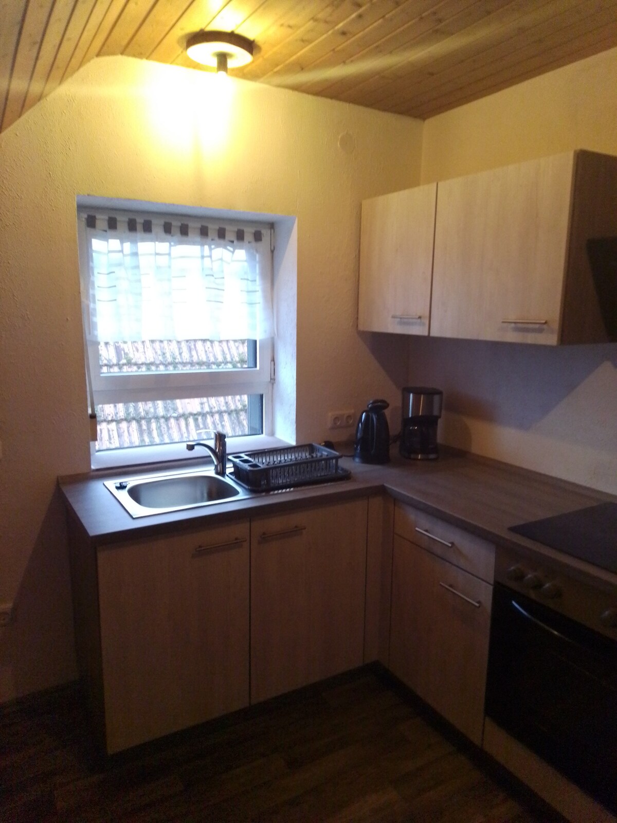 The kitchen area features light wooden cabinetry and a neutral countertop. A window with sheer curtains allows natural light to illuminate the space. Essential appliances such as a coffee maker and kettle are present, along with a sink and a stove for cooking needs.