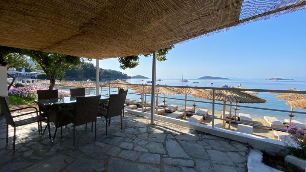 A shaded outdoor seating area features a dining table surrounded by chairs, overlooking the calm sea. Sun loungers are arranged under straw umbrellas on the sandy beach. The scene includes vibrant flowers and distant islands in the background, complemented by a clear blue sky.