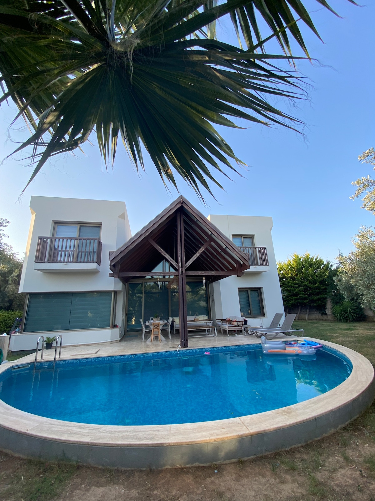 A modern villa features a dual-level design with a distinct roof overhang, surrounded by lush greenery. A swimming pool is positioned prominently in the foreground, with sun loungers and a small table nearby. The facade showcases large glass doors inviting natural light.