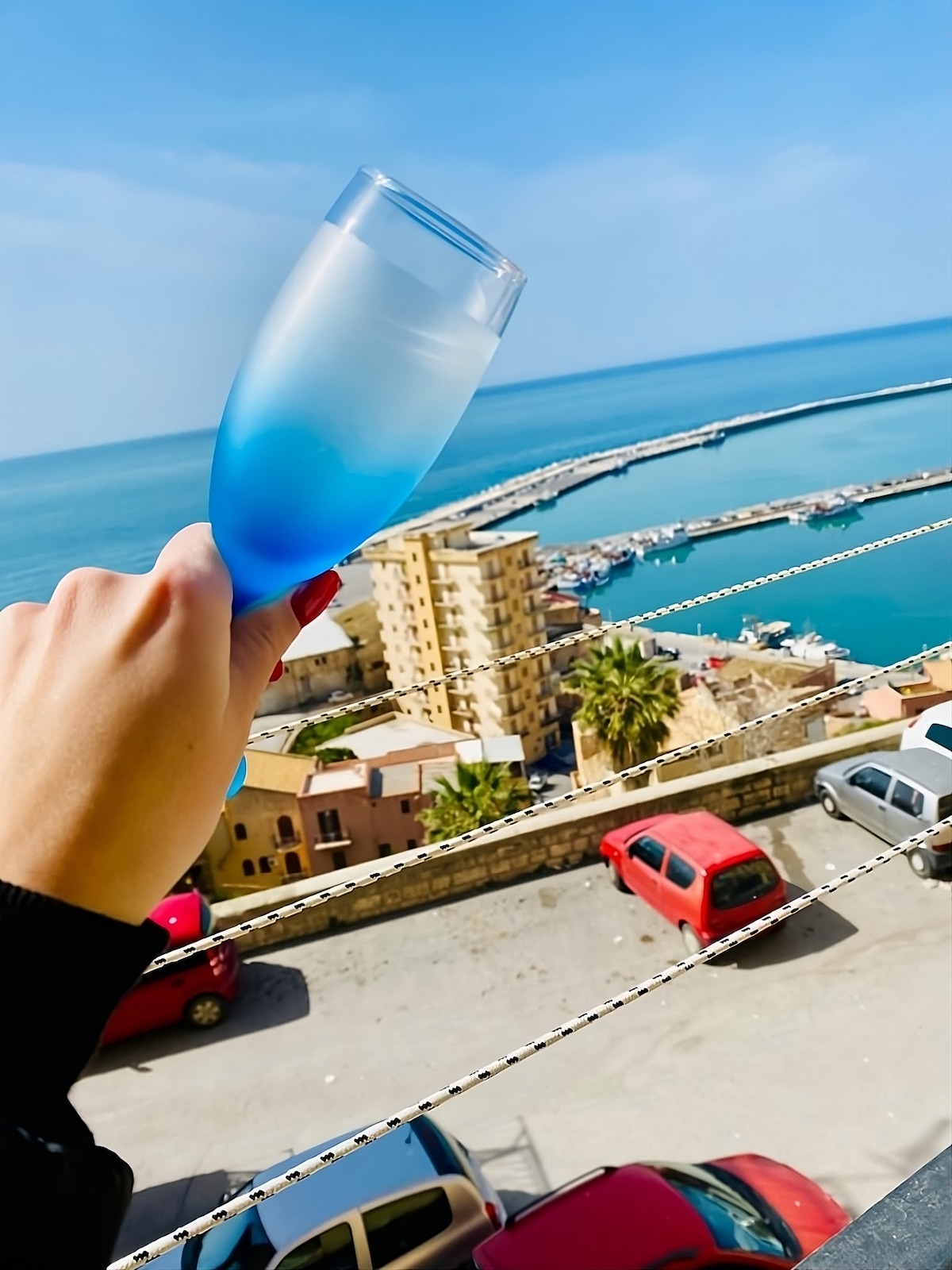 A hand holds a glass with a gradient blue drink, overlooking the marina and sea. The view includes buildings and parked cars along the shoreline, with a clear sky above and hints of coastal landscape in the distance.