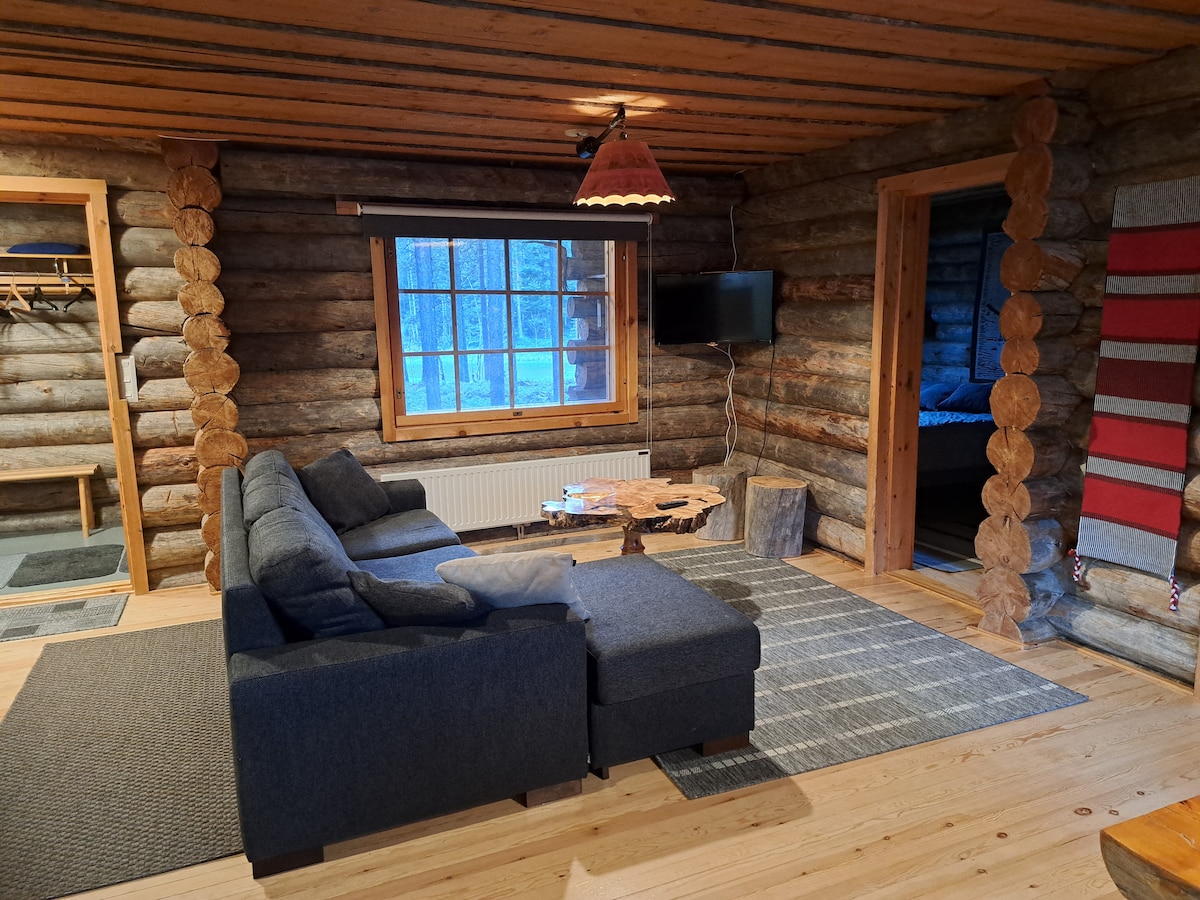 A spacious living area features a comfortable dark gray sectional sofa paired with a rustic wooden coffee table. Natural light fills the room through a large window, while a flat-screen TV is mounted on the wall. Wooden beams and a warm color palette enhance the cozy atmosphere.