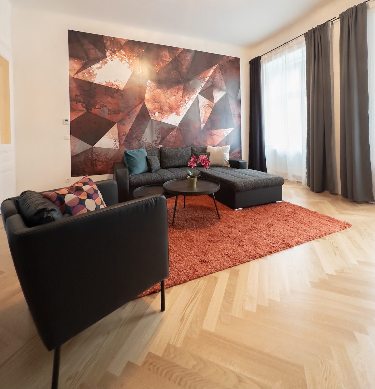 The living area features a deep L-shaped sofa, which complements a single armchair. A round coffee table sits in the center, adorned with a small vase of flowers. A vibrant wall design and large windows introduce natural light, while a textured rug adds warmth to the wooden floor.
