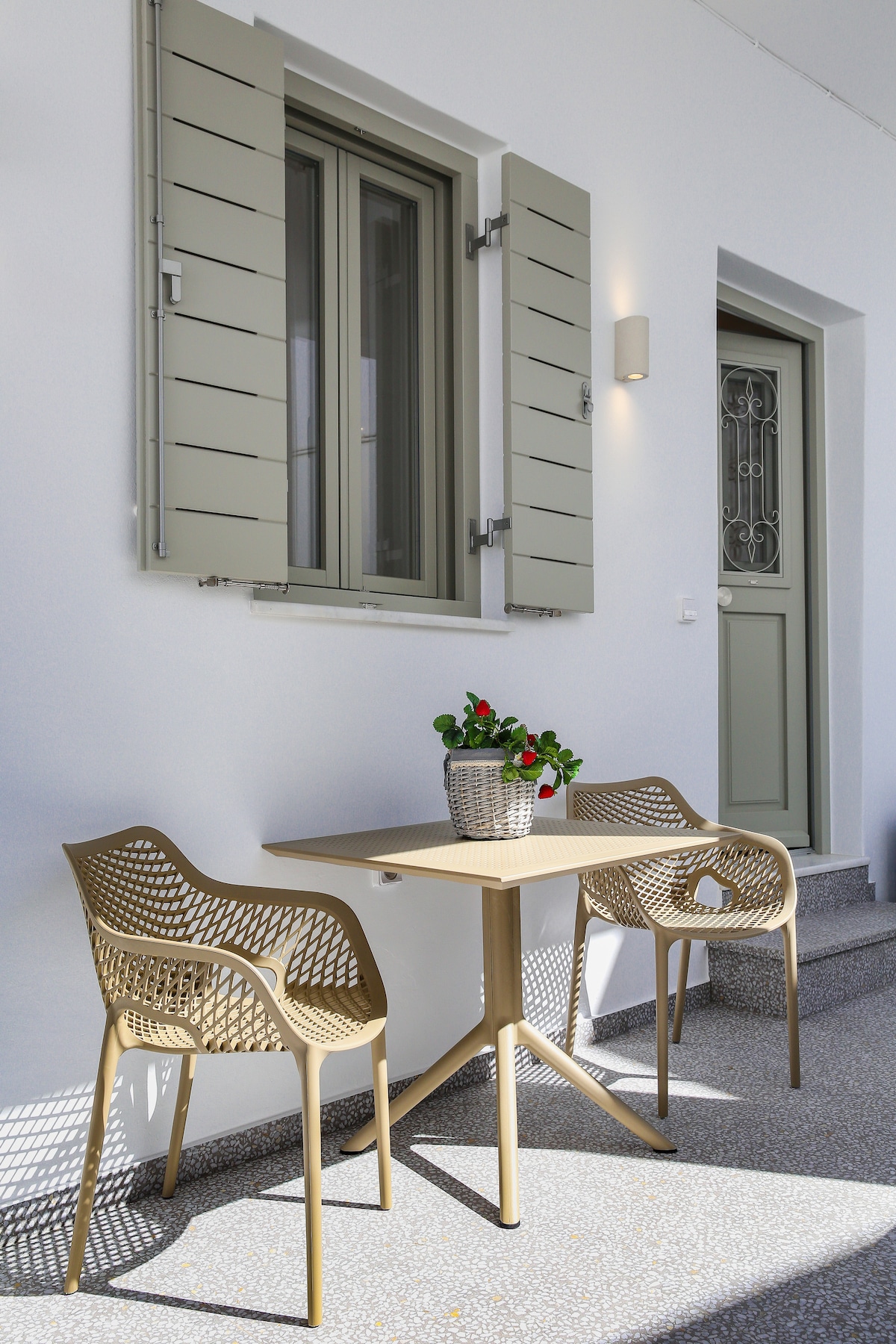 An outdoor seating area features a small table accompanied by two textured chairs. A decorative basket with vibrant flowers rests on the table. The wall behind showcases a window with modern shutters, providing a welcoming entrance to the nearby door.