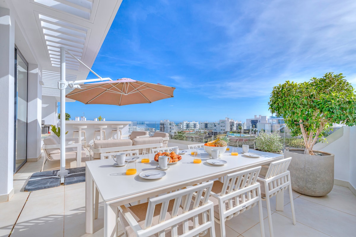 An outdoor dining area features a spacious white table set with plates and bowls. An umbrella provides shade, and a vibrant view of the surrounding area is visible. Comfortable seating and a decorative plant enhance the inviting atmosphere of the terrace.