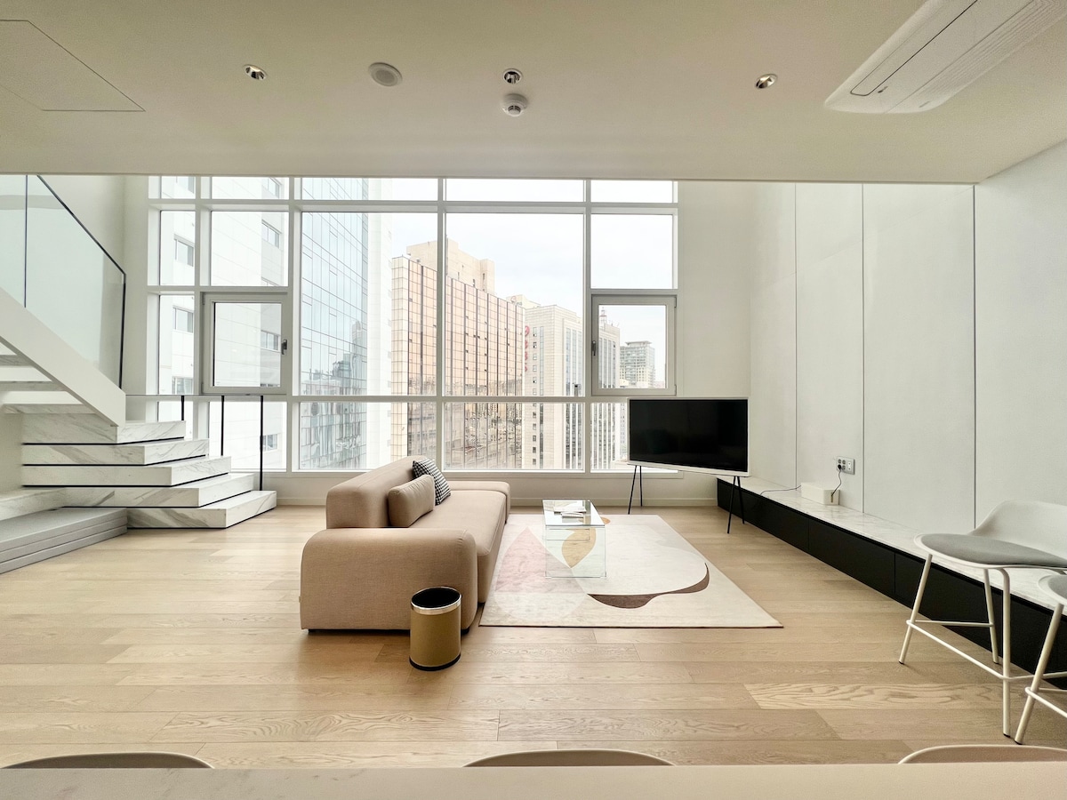 A spacious living area features large windows that offer urban views. A light-colored sofa is positioned centrally on a patterned rug, while a television is mounted on the wall. Minimalist stairs lead to the upper level, and simple furniture arrangements enhance the open layout.