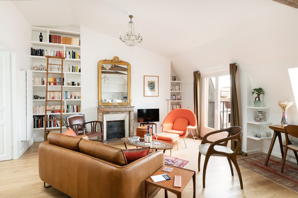 The living room features a comfortable leather couch, an orange accent chair, and a wooden table. A decorative fireplace adds character, and a large mirror reflects natural light. Bookshelves filled with books line one wall, while windows provide views of the surrounding area.