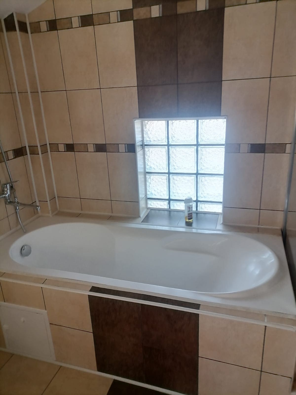 A spacious bathtub is set against a tiled wall, featuring a combination of light and dark ceramic tiles. A small window with glass blocks allows natural light to illuminate the area. A bottle is positioned on the ledge near the window.