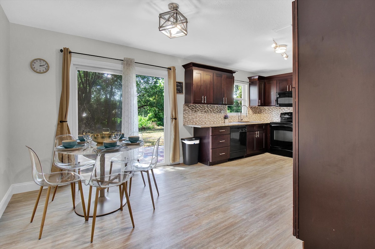 A modern kitchen is visible, featuring dark wooden cabinets and stainless steel appliances. A round glass dining table is set with dishware, surrounded by clear acrylic chairs. Natural light streams through large sliding glass doors that lead to a view of the outdoor patio.