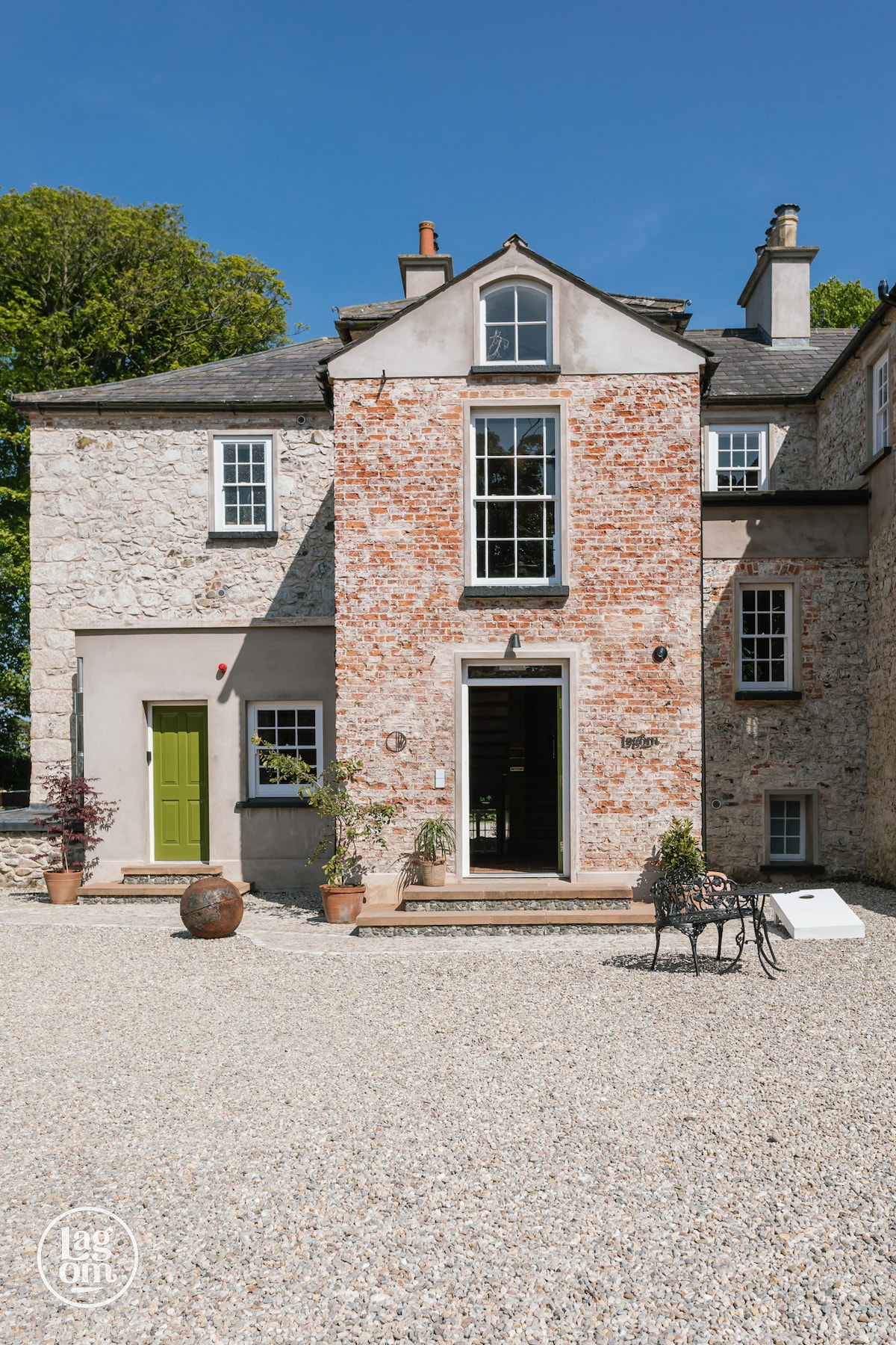 The exterior of the house features a blend of stone and brick, with a welcoming green door at the center. Sunlight illuminates the facade, accentuating the textures. Potted plants add greenery to the gravel driveway, which provides ample space for parking.