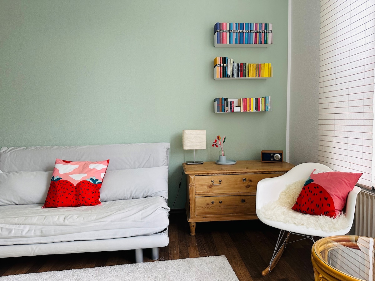 A comfortable seating area features a light green wall and a soft, light-colored rug. A folding sofa with decorative pillows complements a wooden side table, which supports a small lamp and a vintage radio. A wall-mounted shelf holds an array of colorful books or decor items.