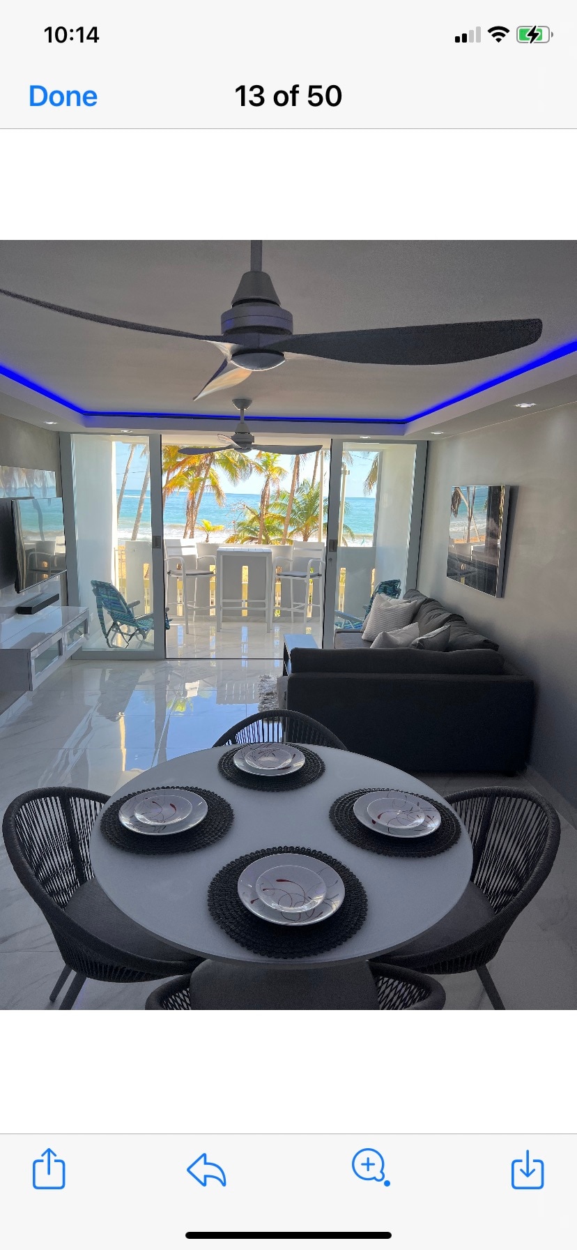 A modern living area features a comfortable sofa and a dining table set for four, presenting a sleek design. Large windows allow natural light to fill the space and offer views of palm trees and the ocean beyond. Blue LED lighting enhances the ambiance.
