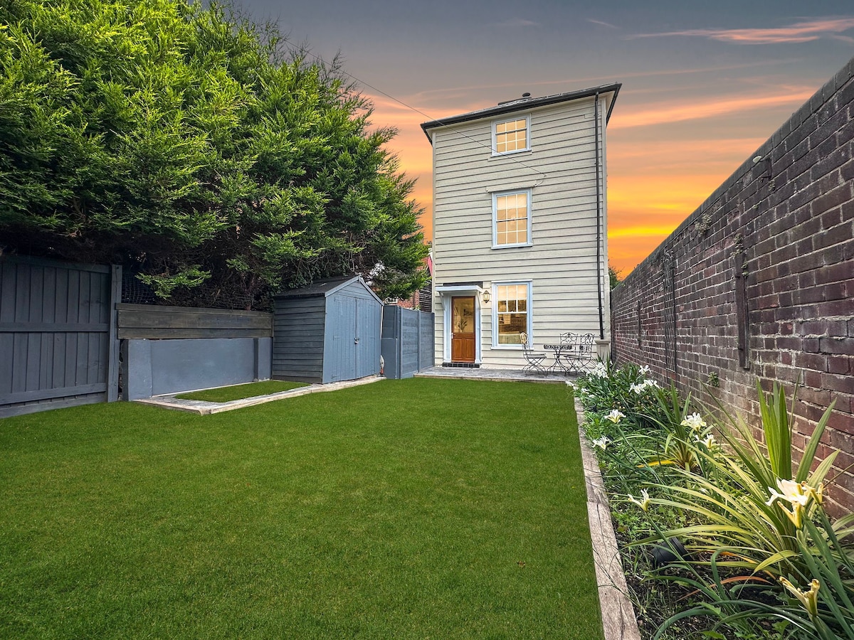 A private walled garden is presented, featuring lush green grass and a pathway lined with flower beds. A two-story wooden cottage stands in the background, with a clear view of its entrance and a garden shed. The sky is illuminated with sunset hues.