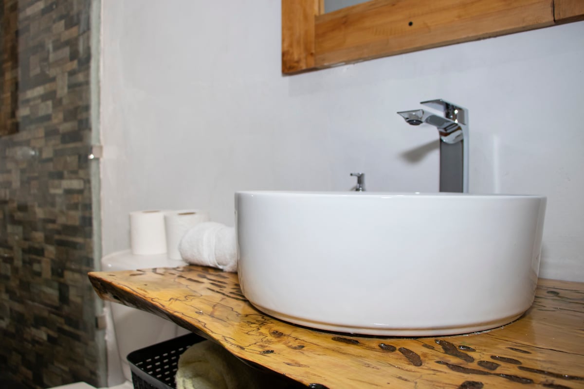 A modern bathroom sink is positioned on a rustic wooden countertop, complemented by a natural wood-framed mirror. White towels are neatly rolled nearby, with toilet paper placed on the side, enhancing the functional and clean spaces offered within the cabin.