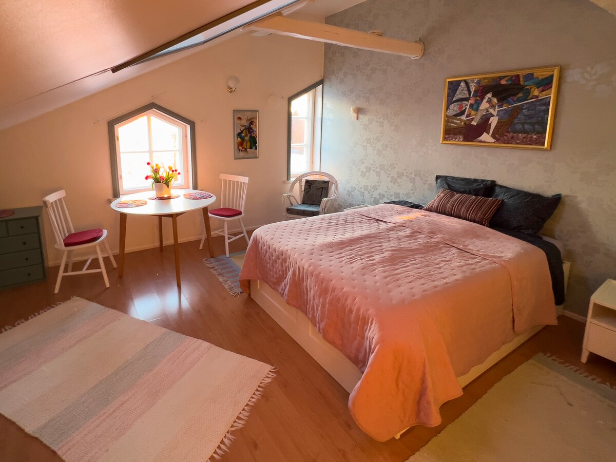 A bright and airy room features a spacious bed with a soft pink coverlet. Two windows allow natural light to fill the space, complemented by a small dining table with two chairs. A decorative rug adds warmth, while artwork adorns the walls.