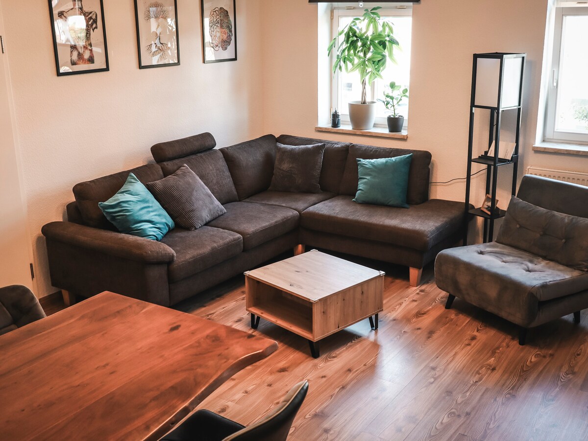 A spacious living area features a large, brown sectional sofa adorned with teal cushions. A small wooden coffee table is positioned in the center, surrounded by a dining table with dark chairs. Potted plants sit by the windows, allowing natural light to fill the room.