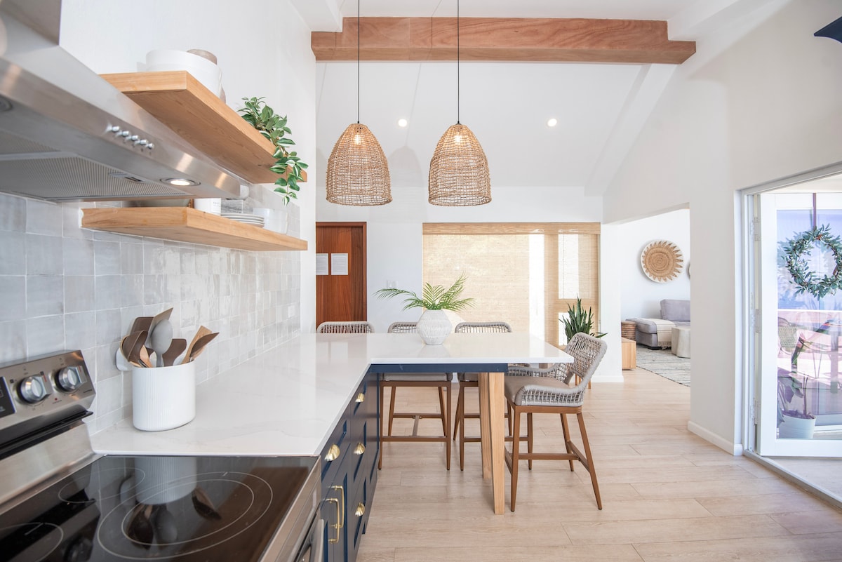 An eat-in kitchen features modern appliances, white countertops, and natural wood accents. Two woven pendant lights hang above a central island with seating. Open shelving displays cooking essentials, while the light-colored walls and flooring contribute to an airy feel. An adjoining living area is visible through the doorway.