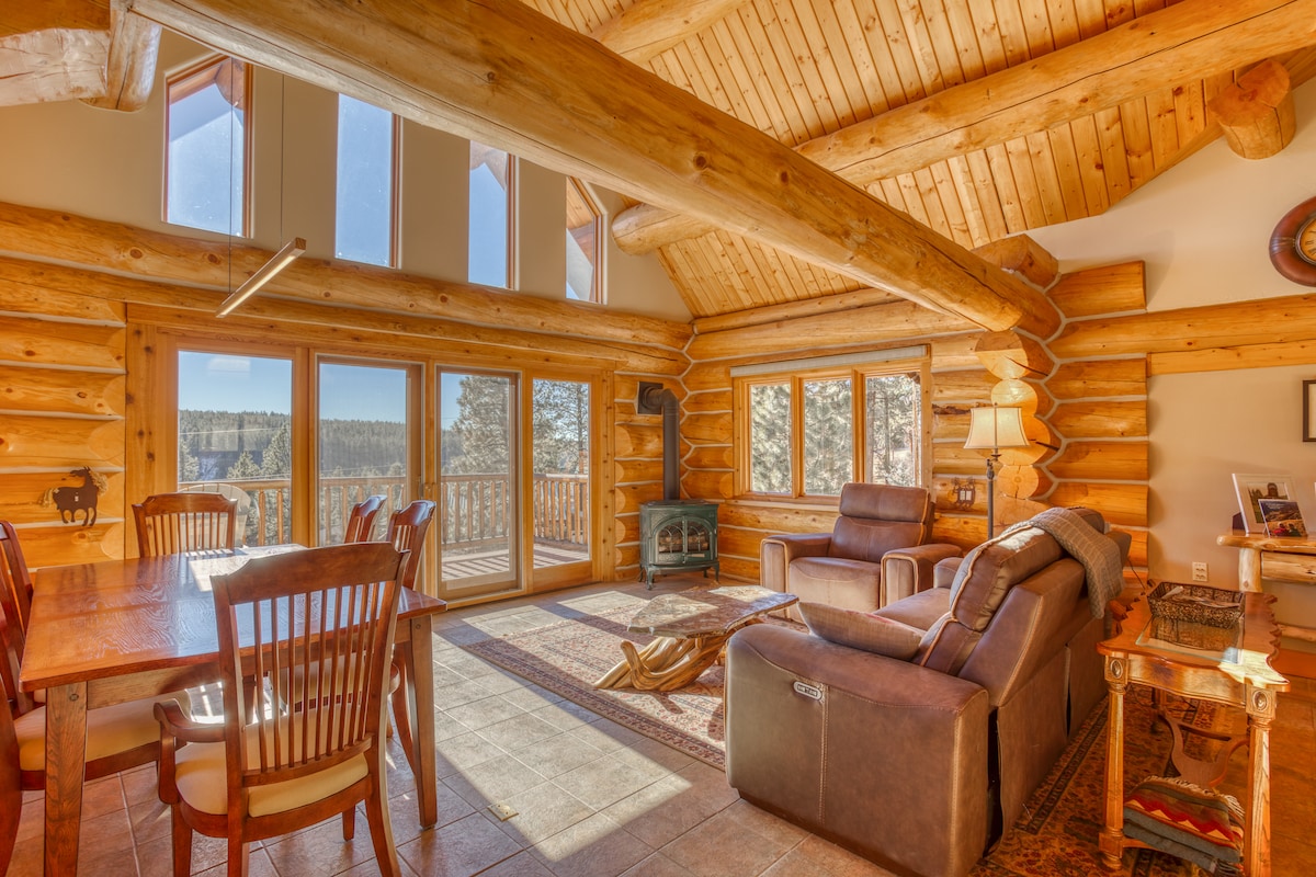 A spacious interior features a combination of log walls and high ceilings, allowing natural light to fill the room. Comfortable seating options are arranged around a decorative rug, with a dining area visible in the background. Large windows offer a view of the surrounding nature.