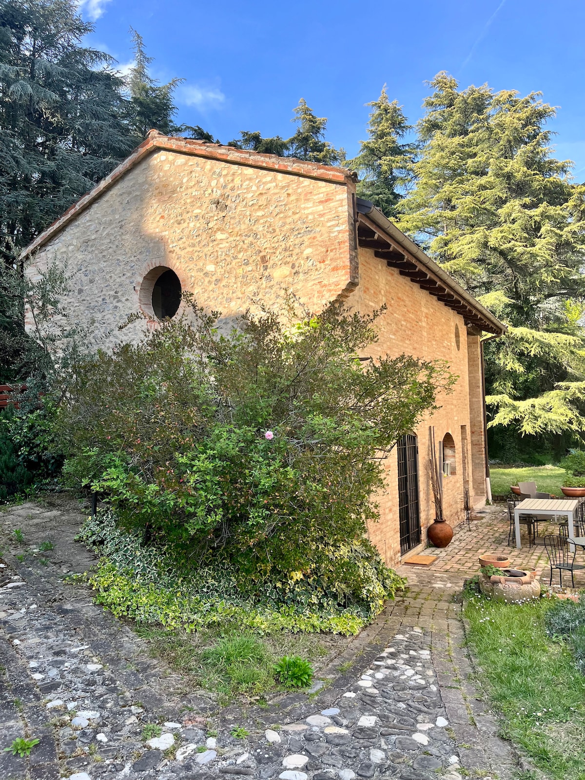 A charming stone building is surrounded by lush greenery, with tall trees and flowering plants framing its facade. A pathway made of stones leads to the entrance, where outdoor seating is visible, offering a serene retreat within the natural landscape.