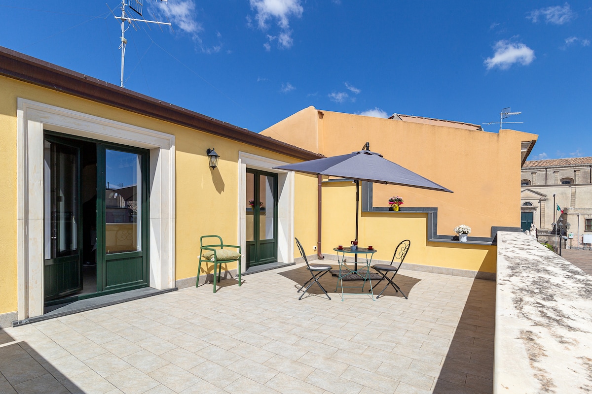 An expansive terrace offers outdoor seating arrangements, including a round table and four chairs shaded by a large umbrella. The light-colored tiled flooring enhances the space, while green doorways lead indoors. Bright colors and a clear blue sky create a cheerful atmosphere.
