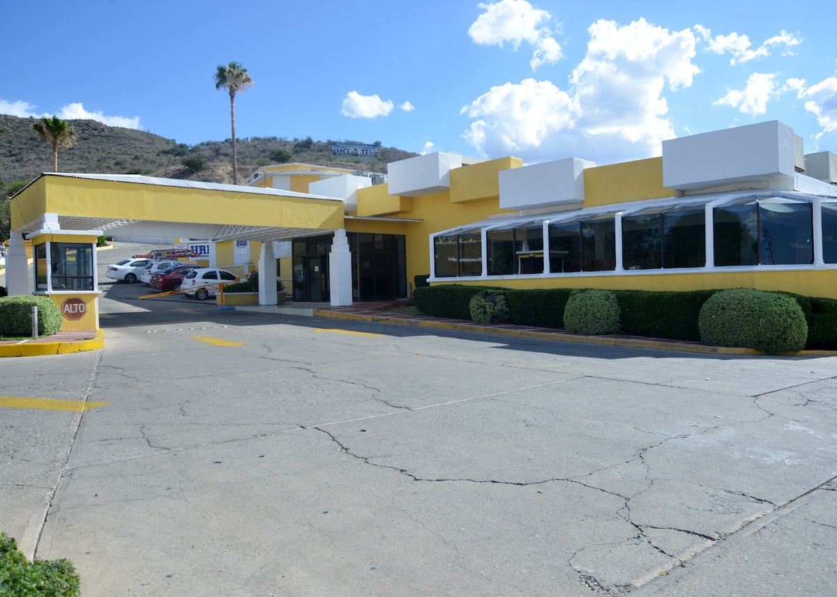 The exterior of a bright yellow building is visible, featuring large windows and a covered entrance. Palm trees are seen in the background against a clear blue sky, while a paved parking area accommodates several vehicles. The surrounding landscape includes gentle hills.