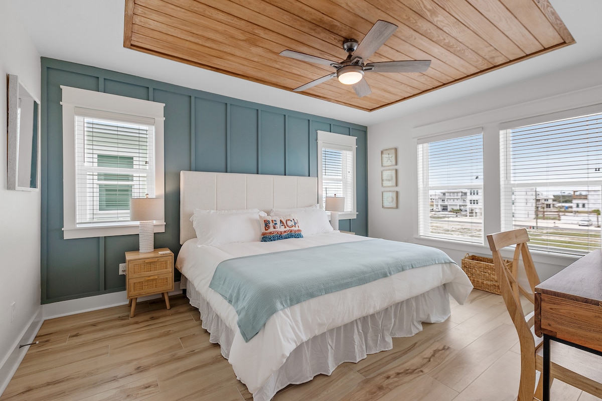 A spacious bedroom features a large bed with a light blue throw blanket, complemented by a decorative pillow. Natural light floods the room through multiple windows with white blinds, and a wooden ceiling adds warmth. A desk and lamp are positioned nearby.