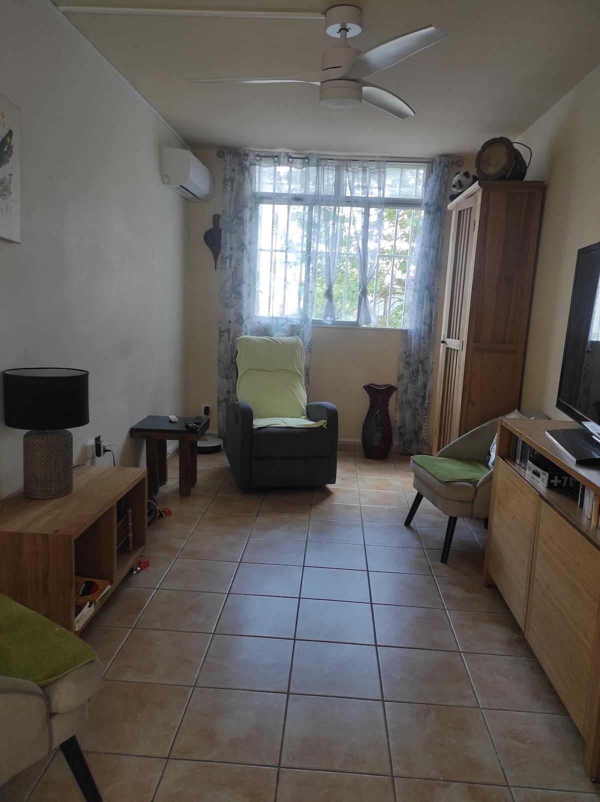 A living space features a comfortable armchair and a small side table, positioned near a window adorned with sheer curtains allowing natural light. A TV unit, a wooden cabinet, and a decorative vase are visible, contributing to a functional and inviting environment.