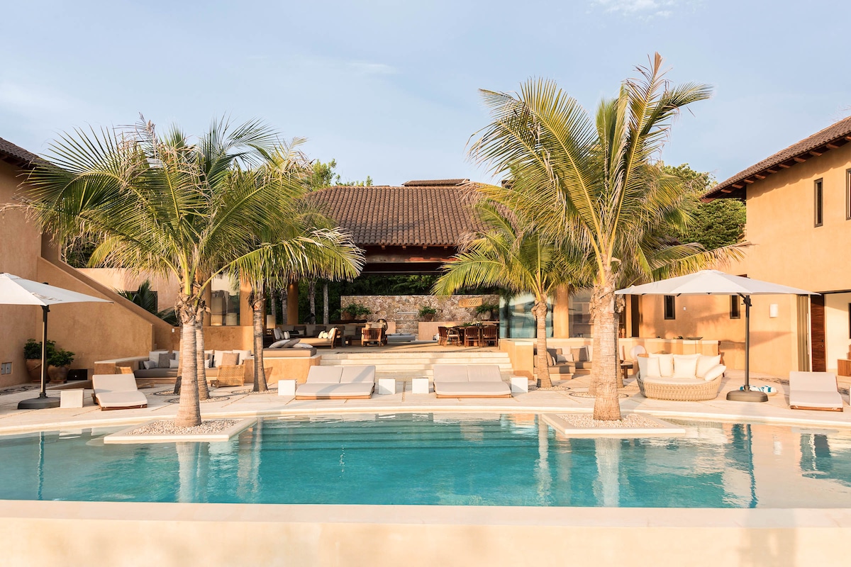 A spacious pool area is framed by palm trees, featuring lounge chairs arranged around the water. An inviting terrace offers additional seating and outdoor dining options, while the villa's warm earth-toned façade complements the serene environment.