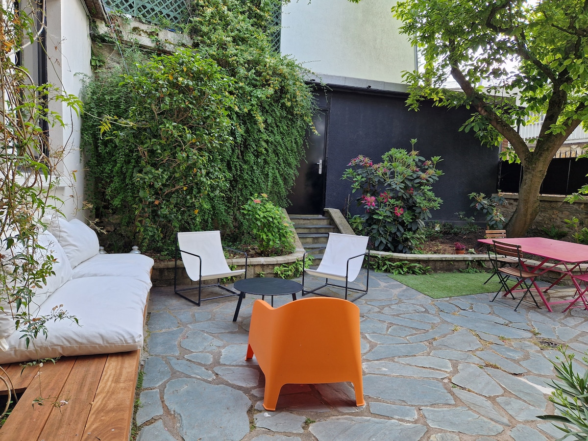 A garden space features a stone patio surrounded by greenery. Comfortable seating is provided, including a white sofa, armchairs, and a round black table. Additional vibrant furnishings are present, with a small grassy area and steps leading to a darker section of the garden.