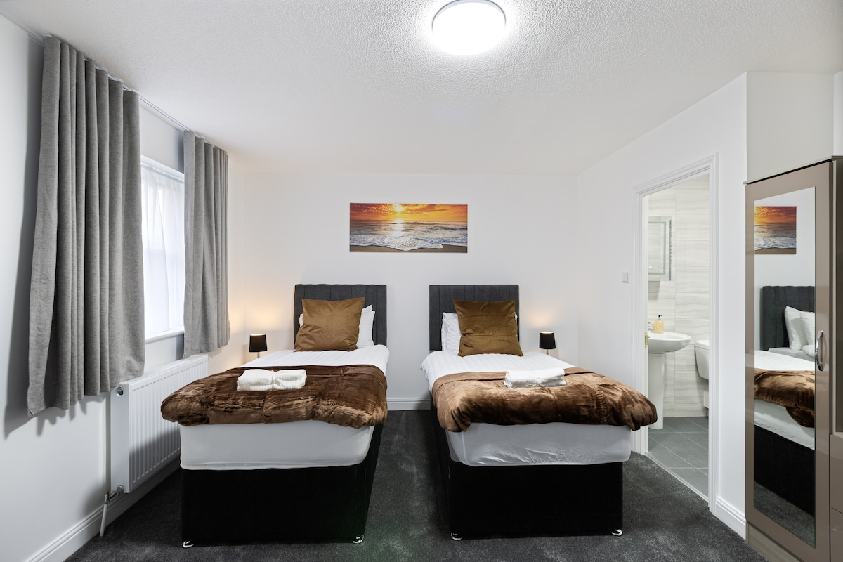 A bright bedroom is furnished with two twin beds, each featuring plush brown bedding. Decorative pillows complement the arrangement. A piece of artwork depicting a serene sunset adorns the wall above the beds. Soft grey curtains frame the window, adding warmth to the space.