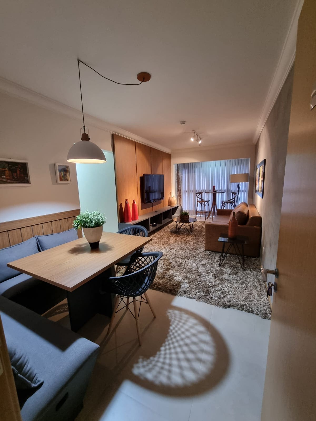 A modern living space is displayed with a dining area featuring a wooden table surrounded by black chairs. A cozy seating area includes a brown sofa and a carpeted floor. The room is brightly lit, enhancing the ambiance with large windows visible in the background.