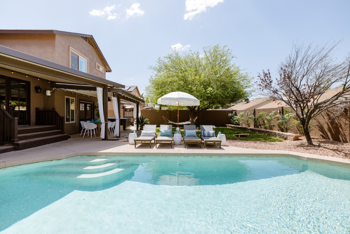A serene outdoor space features a sparkling pool surrounded by lounge chairs. An umbrella provides shade, while the home’s expansive patio offers seating for relaxation. Lush greenery and landscaped areas enhance the tranquil atmosphere under a clear blue sky.