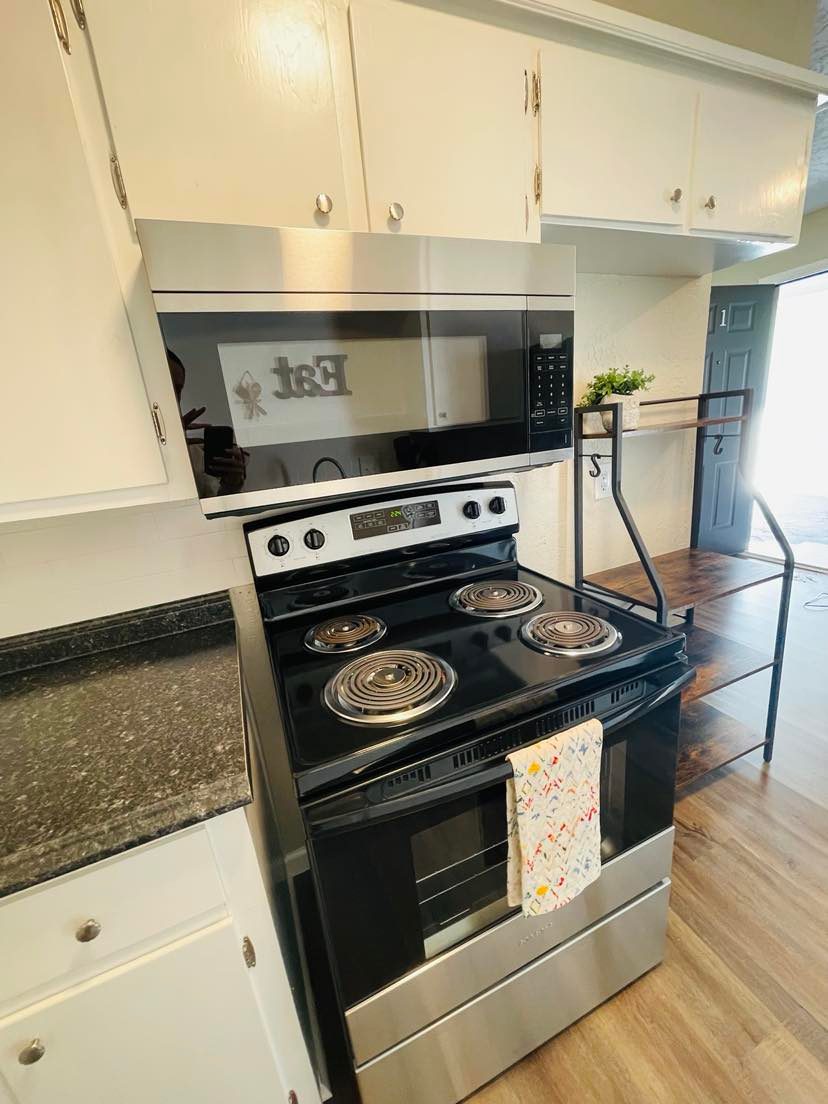 A modern kitchen features stainless steel appliances, including a sleek stove and microwave. The dark countertops contrast with white cabinetry. A decorative towel hangs from the stove, adding a touch of color to the space. A door leading outside is visible in the background.