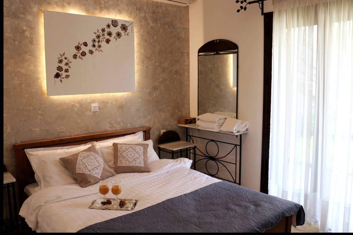 A welcoming bedroom features a spacious bed adorned with neutral-toned linens and decorative pillows. A framed floral artwork is displayed above the bed, complemented by soft ambient lighting. Natural light floods the room through sheer curtains, with a mirror and neatly arranged towels visible in the background.