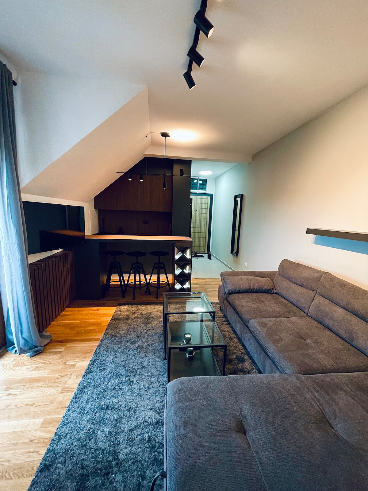 A modern living area features a spacious gray sofa positioned on a plush area rug. A glass coffee table sits in front, while sleek bar stools line a dark wood counter in the background. Soft lighting enhances the open and airy ambiance.