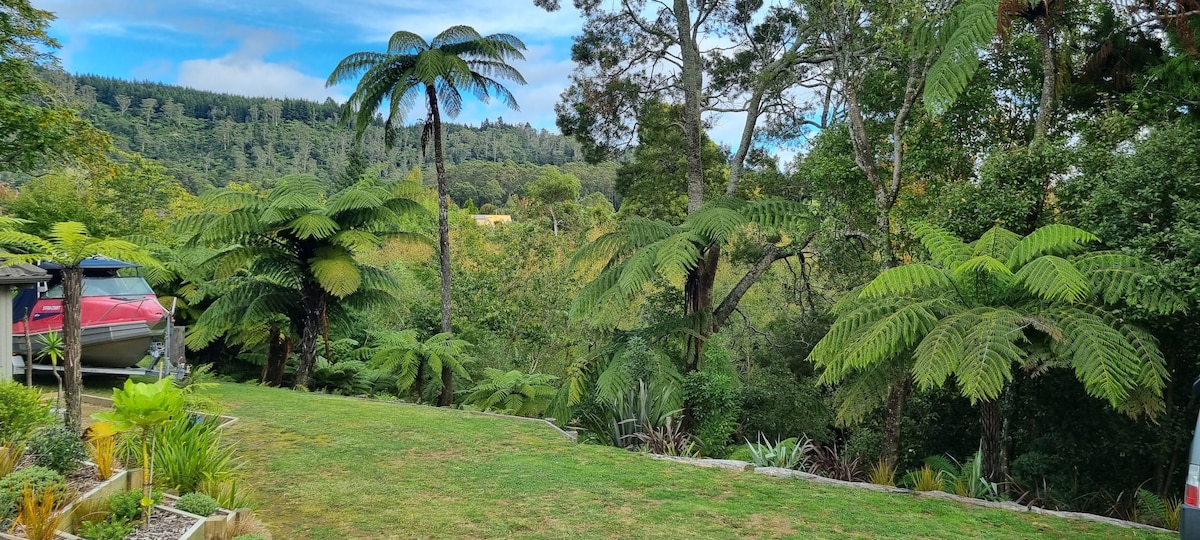 A lush, green outdoor space is surrounded by tall ferns and trees. The expansive lawn is complemented by various plants and a glimpse of a distant hillside, creating a serene natural setting.